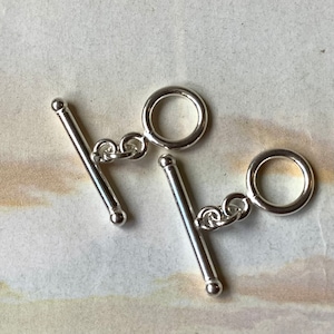May include: Two silver-colored toggle clasps. Each clasp features a T-bar and a ring connected by a small chain. The clasps are displayed against a light-colored, textured background.
