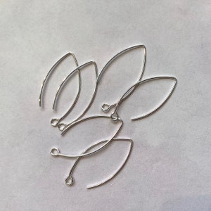 May include: A collection of silver earring hooks, each with a curved, elongated design and a small loop, are arranged on a white background. The hooks are made of a shiny, reflective metal.
