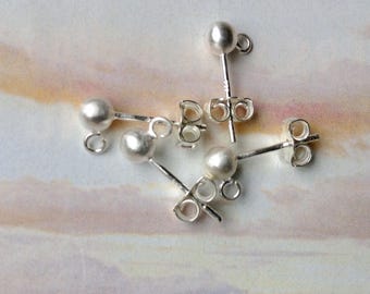 May include: A collection of silver-colored ball stud earrings with posts and butterfly backs. The earrings are scattered on a light-colored surface with a soft, blurred background. The earrings are small and delicate, suitable for everyday wear.