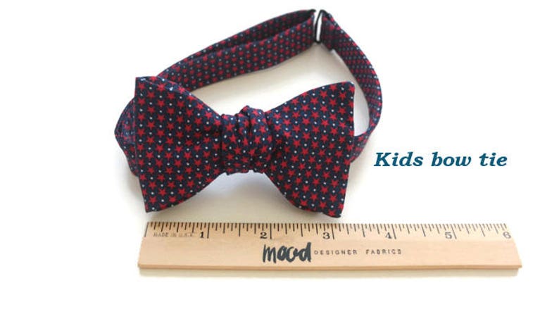 May include: A red and navy blue bow tie with a white star pattern. The bow tie is tied and ready to wear. A ruler is in the image for size reference. "Kids bow tie" is written in blue text on a white background.