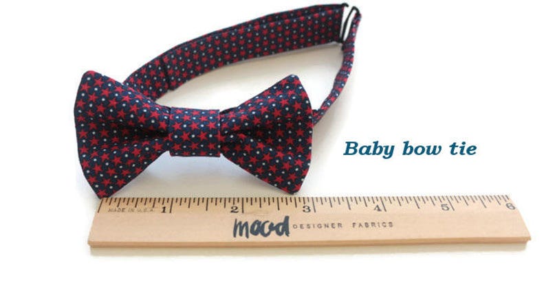 May include: A navy blue bow tie with red stars, measuring approximately 3 inches in length. The bow tie is made of fabric and has an adjustable strap. The text "Baby bow tie" is visible in the image.