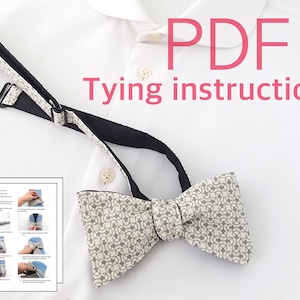 May include: A white bow tie with a gray geometric pattern and black adjustable strap. The bow tie is on a white surface with a white shirt and a sheet of paper with instructions on how to tie the bow tie. The paper has the text "PDF Tying instruction".