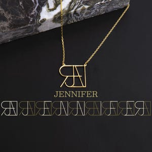 May include: A gold necklace with a geometric design featuring the letters "RJ" in a square shape. The necklace is on a black background with the word "JENNIFER" written below the pendant.