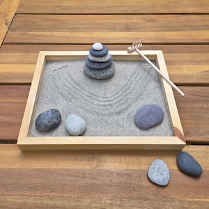 May include: A small zen garden with a wooden frame, filled with gray sand. A stack of gray stones is centered, with a small wooden rake and several loose stones. The sand has a raked pattern.