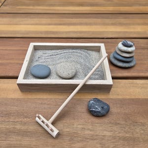 May include: A miniature Zen garden set with a wooden tray filled with sand, two smooth stones, and a small wooden rake. A stack of stones and a single stone are also present, all on a wooden surface.