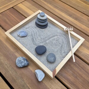 May include: A small zen garden with a light wood frame, gray sand, and smooth gray stones. A stack of stones and a small wooden rake are also included. The garden sits on a wooden surface.