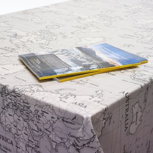May include: A white tablecloth with a black and white world map print. Two travel magazines are stacked on top of the tablecloth. The top magazine has the text "TRAVEL BEYOND YOUR NEXT GREAT ADVENTURE" on the cover.