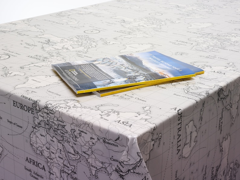May include: A white tablecloth with a black and white world map print. Two travel magazines are stacked on top of the tablecloth. The top magazine has the text "TRAVEL BEYOND YOUR NEXT GREAT ADVENTURE" on the cover.