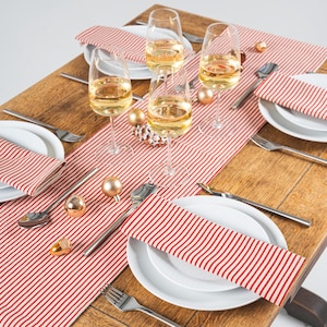 May include: A Christmas table setting featuring white plates, red and white striped napkins, and champagne flutes filled with a golden liquid. Silver cutlery and gold baubles are arranged on a wooden table, creating a festive atmosphere.