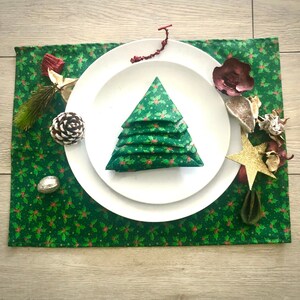 May include: A Christmas-themed table setting with a green holly berry patterned placemat. White plates support a folded napkin in the shape of a Christmas tree. Decorations include a pine cone, star, and dried botanicals.
