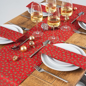 May include: A Christmas table setting featuring a red runner and napkins with a green holly design. Wine glasses with a golden liquid, white plates, and silver cutlery are arranged on a wooden table. Gold baubles and a pine cone add festive detail.