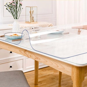 May include: A clear plastic table protector is placed on top of a wooden table. The table protector is designed to protect the table surface from scratches and spills. The table protector is clear and has a smooth surface.