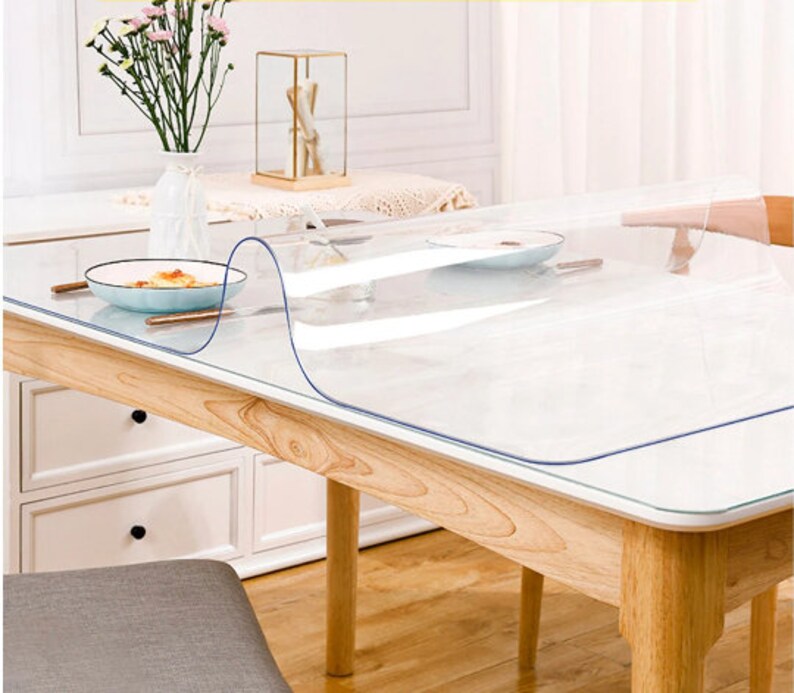 May include: A clear plastic table protector is placed on top of a wooden table. The table protector is designed to protect the table surface from scratches and spills. The table protector is clear and has a smooth surface.