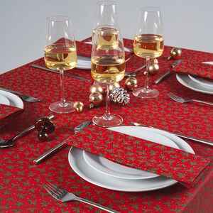 May include: A Christmas-themed table setting with a red tablecloth patterned with green holly. White plates, silverware, and wine glasses filled with a golden liquid are arranged. Gold baubles and a decorative stopper complete the festive look.