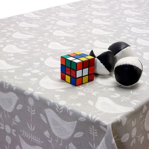 May include: A grey tablecloth featuring a white bird and floral design. A multi-coloured Rubik's Cube and three black and white juggling balls are on the cloth. The tablecloth is draped over a table.