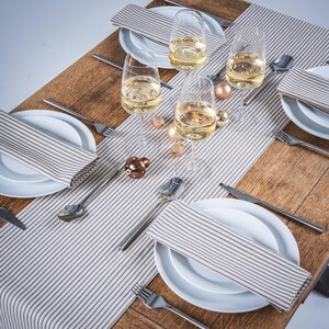May include: A dining table set with white plates, striped napkins, silverware, and wine glasses filled with a light-coloured liquid. Decorative gold and brown baubles are scattered on the table runner.
