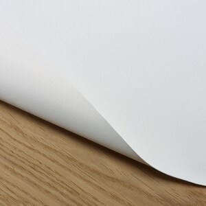 Puede incluir: Primer plano de una tela de lona blanca con un ligero pliegue, mostrando la textura del material. La tela est&aacute; sobre una superficie de madera marr&oacute;n claro.