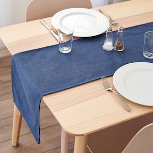 May include: A navy blue table runner on a light wood table, set with white plates, clear glasses, and cutlery. Salt and pepper shakers are also on the table. The table is set for a meal.