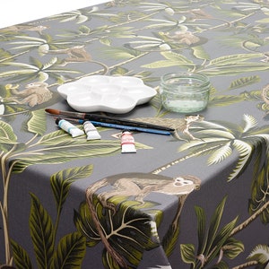 May include: A grey tablecloth with a jungle print featuring monkeys and palm trees. The tablecloth has paintbrushes, paint tubes, a palette, and a glass jar on top of it.