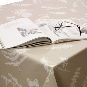 May include: A beige tablecloth with a white botanical design of ferns, flowers, and butterflies. An open book and black-framed glasses are on the cloth, creating a calm, nature-inspired scene.