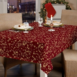 May include: A rectangular burgundy tablecloth with a gold leaf pattern covers a table set for a meal. The table is set with plates, glasses, and silverware. A lit candle sits in a decorative holder.