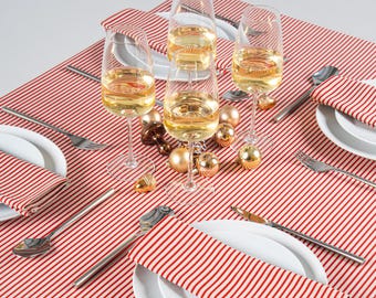 May include: A Christmas table setting with a red and white striped tablecloth. White plates, silverware, and red and white striped napkins are arranged. Wine glasses filled with a golden liquid and gold baubles complete the festive scene.