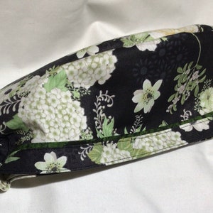 May include: Black scrub hat with a white and green floral print. The hat has a ruffle at the back and a green trim around the edge.