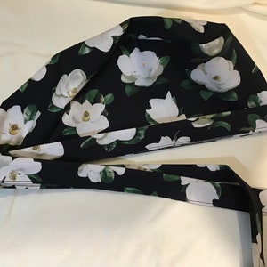 以下が含まれることがあります： 白いマグノリアの花柄の黒い布製のスクラブキャップ。帽子には、頭の後ろで結ぶことができるリボンが付いています。