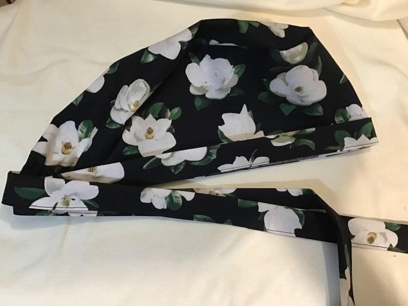 以下が含まれることがあります： 白いマグノリアの花柄の黒い布製のスクラブキャップ。帽子には、頭の後ろで結ぶことができるリボンが付いています。