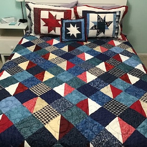 May include: A blue, red, and white patchwork quilt with a geometric pattern. The quilt is on a bed with two pillows with stars on them.