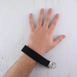 May include: A black woven wristlet keychain with a silver key ring. The keychain is worn on a person's wrist.