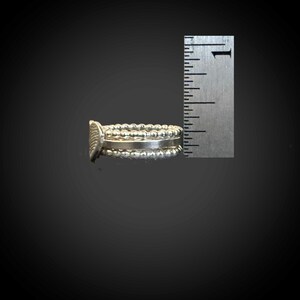 May include: A silver ring with a textured design and beaded accents. The ring is shown next to a ruler, with a measurement of approximately 0.75 inches. The ring has a detailed, ornate appearance.