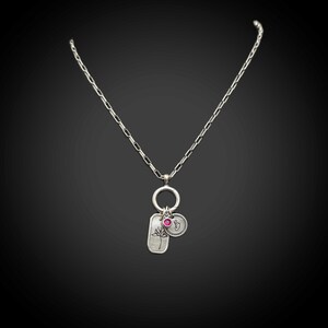 May include: Silver necklace with a rectangular pendant featuring a tree design, a circular charm with the letter "b", and a small pink gemstone. The necklace has a delicate chain and a round clasp.