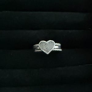 May include: A silver ring featuring a heart-shaped design with a fingerprint pattern. The ring has a double band with a twisted rope texture. The ring is set against a black background.