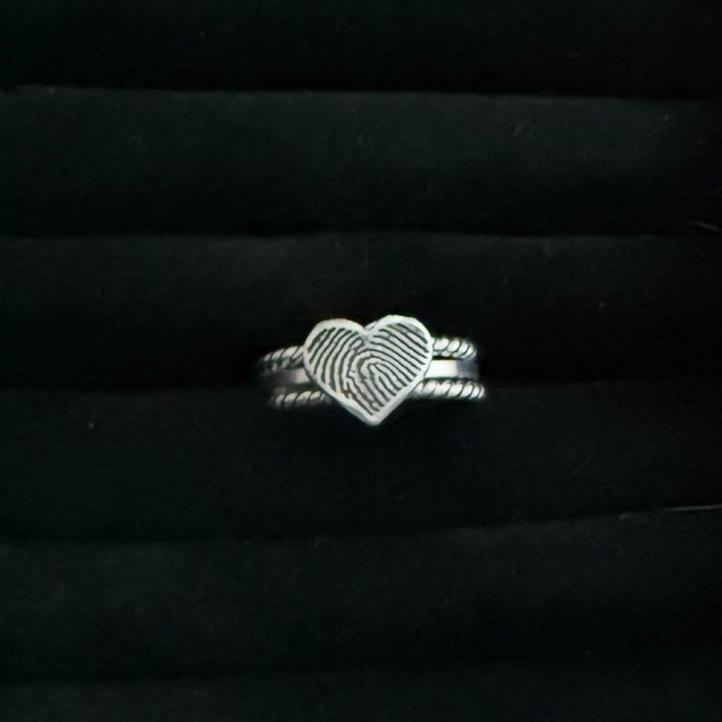 May include: A silver ring featuring a heart-shaped design with a fingerprint pattern. The ring has a double band with a twisted rope texture. The ring is set against a black background.