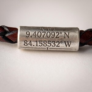 May include: A bracelet featuring a cylindrical silver-toned metal bead with a weathered finish. The bead is engraved with the coordinates 9.407092 N and 84.158532 W. The bead is threaded onto a braided dark brown leather cord that extends on both sides.