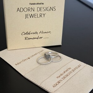 May include: A set of silver rings with a heart design and beaded band, displayed on a beige fabric. The rings are next to a box and fabric with the text "Handcrafted by ADORN DESIGNS JEWELRY" and "Celebrate, Honor, Remember".