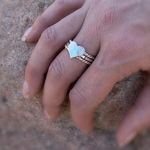 May include: A silver ring set featuring a heart-shaped centerpiece. The ring is composed of three bands, with the top band showcasing the heart design. The other two bands have a twisted rope design. The ring is worn on a finger.