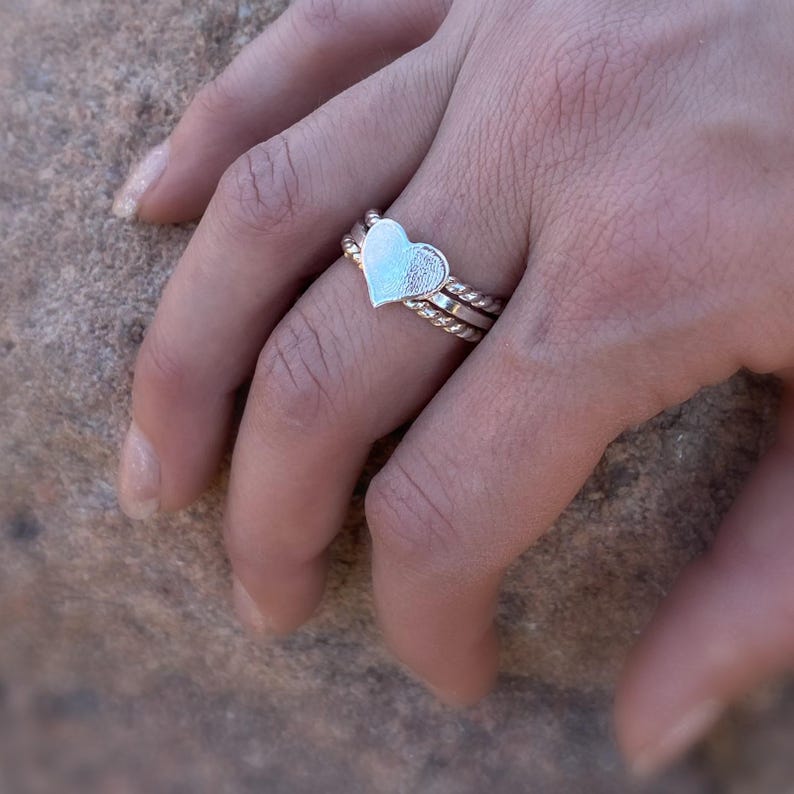 May include: A silver ring set featuring a heart-shaped centerpiece. The ring is composed of three bands, with the top band showcasing the heart design. The other two bands have a twisted rope design. The ring is worn on a finger.