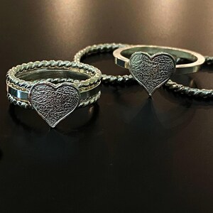 May include: Two silver rings with heart-shaped fingerprint designs. One ring has a braided band, while the other has a simple band. The rings are displayed on a dark surface, showcasing the intricate details of the fingerprint impressions.