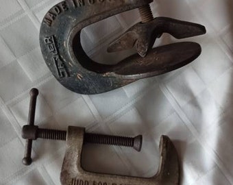 May include: Two vintage metal C-clamps. The top clamp is dark, marked "SHAEFER" and "MADE IN USA" with a wing nut screw. The bottom clamp is lighter, marked "JUDD 500 POUND TEST" and features a T-handle screw mechanism for tightening the clamp.