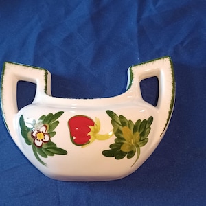 May include: A white ceramic vase with two handles and a strawberry design. The vase has a curved shape and is decorated with a red strawberry, green leaves, and a white flower with yellow and red accents. The handles are outlined in green.