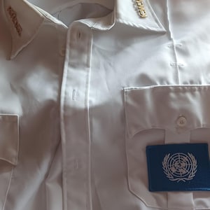 May include: White button-down shirt with a collar and two chest pockets. Features gold-colored pins on the collar and a blue patch with a white emblem. The shirt has the text "First Class" on the label.