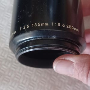 Puede incluir: Parasol de lente negro con el texto "1:3.5 135mm 1:5.6 200mm" impreso en el lateral.