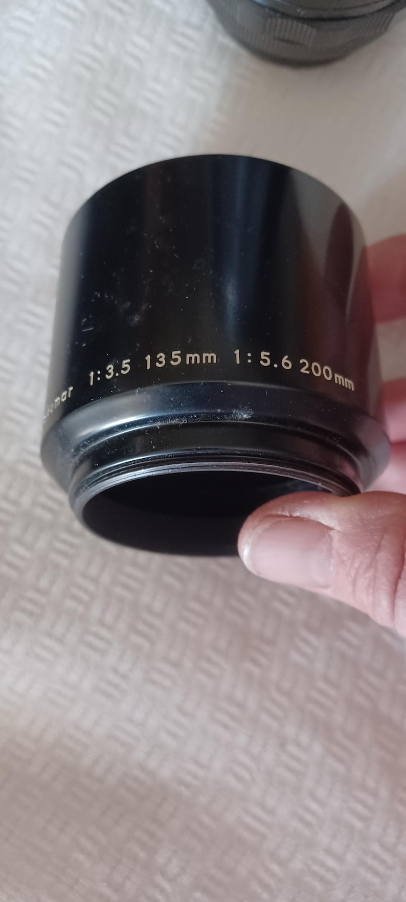 Puede incluir: Parasol de lente negro con el texto "1:3.5 135mm 1:5.6 200mm" impreso en el lateral.