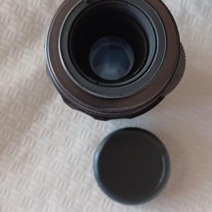 Puede incluir: Un objetivo de c&aacute;mara negro con un anillo plateado alrededor del borde. El objetivo est&aacute; sobre una superficie blanca con una tapa de objetivo negra al lado.