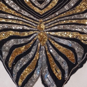 Puede incluir: Prenda negra con forma de mariposa adornada con lentejuelas doradas y plateadas. La prenda est&aacute; colgada de una percha negra. Las lentejuelas est&aacute;n dispuestas en un patr&oacute;n radial, creando un efecto visual llamativo.