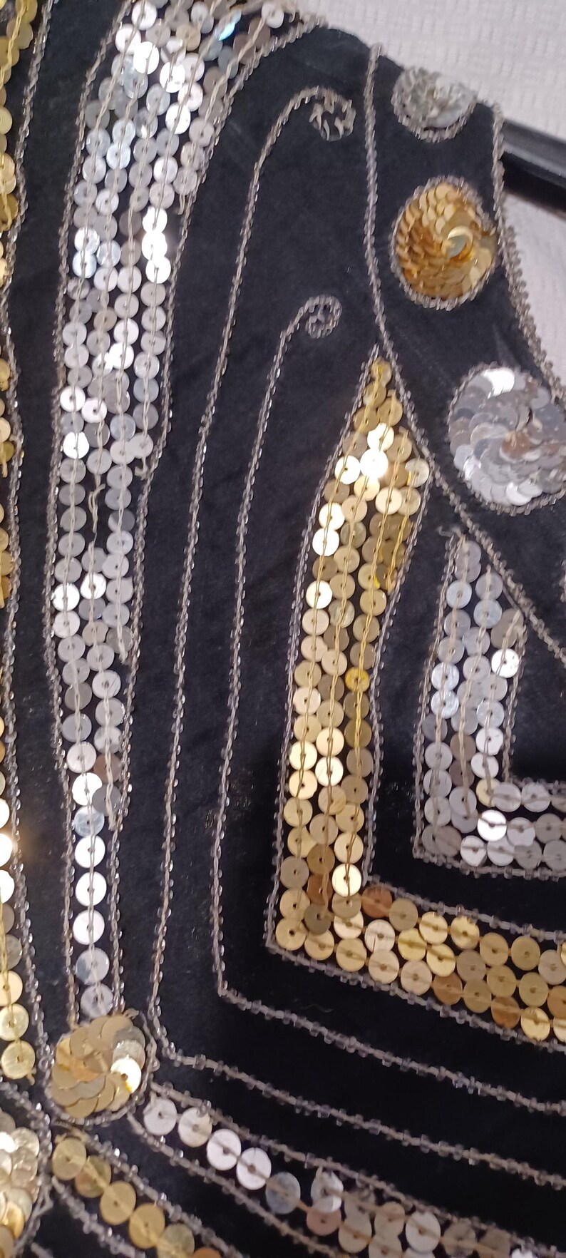 Puede incluir: Primer plano de una prenda negra adornada con lentejuelas plateadas y doradas. Las lentejuelas est&aacute;n dispuestas en patrones verticales y geom&eacute;tricos, creando un efecto brillante. El dise&ntilde;o sugiere un estilo vintage o formal.