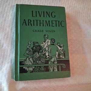 May include: A green hardcover textbook titled Living Arithmetic Grade Seven. The cover features a dark green illustration of a person at a cash register with a customer holding a shopping basket, rendered in a vintage graphic style with black ink.