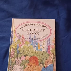 May include: A children's book titled "Little Grey Rabbit's Alphabet Book" by Alison Uttley & Margaret Tempest. The cover features illustrations of rabbits and other animals in a garden setting with colorful flowers. The book is on a blue background.
